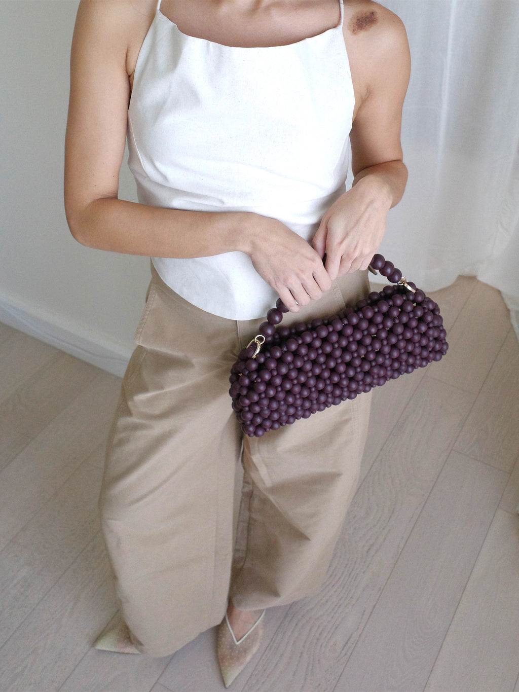 A person wearing a white sleeveless top and beige wide-leg pants holds a burgundy beaded handbag with a matching beaded top handle in a bright indoor setting with light wood flooring.