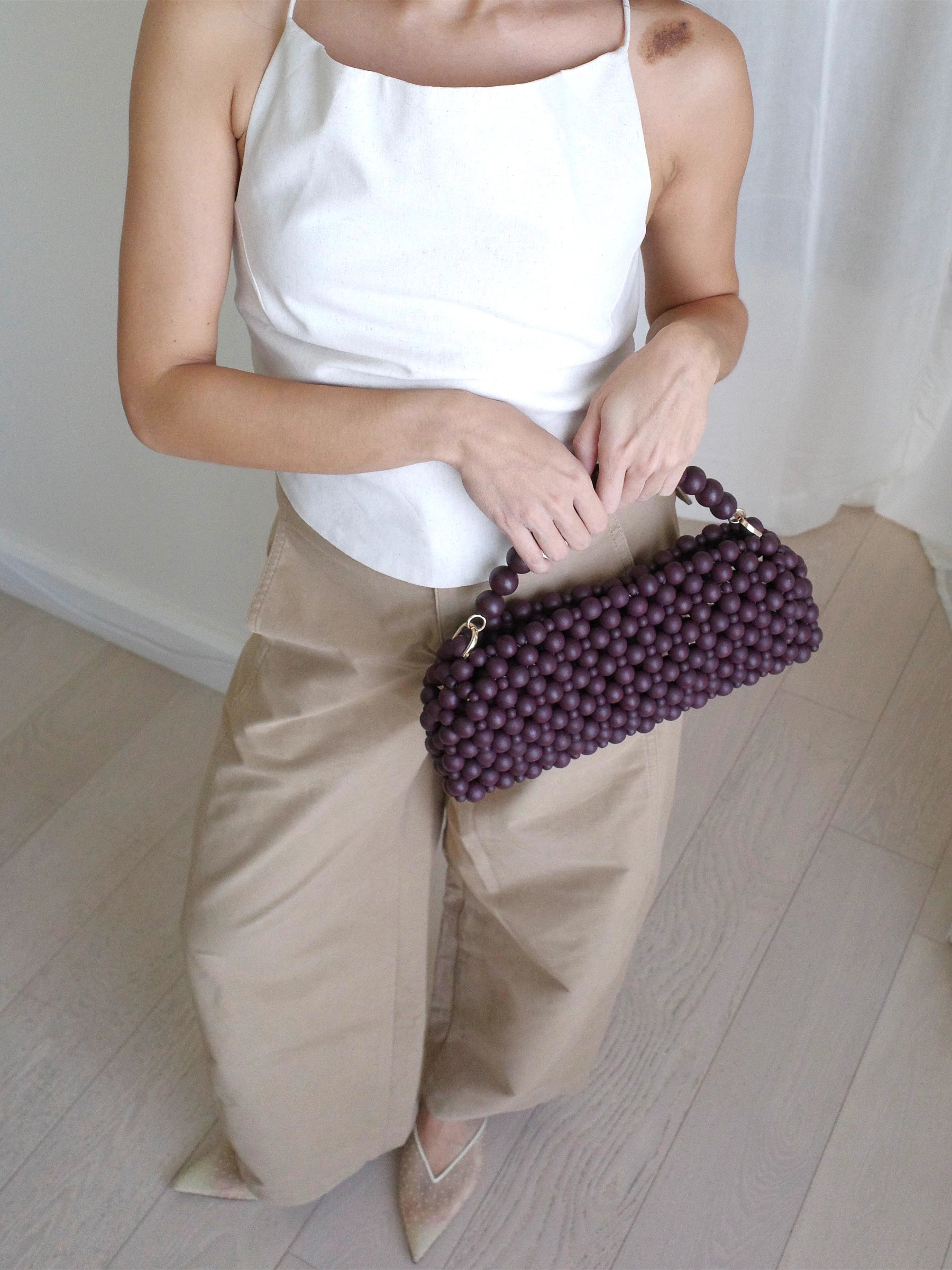 A person wearing a white sleeveless top and beige wide-leg pants holds a burgundy beaded handbag with a matching beaded top handle in a bright indoor setting with light wood flooring.