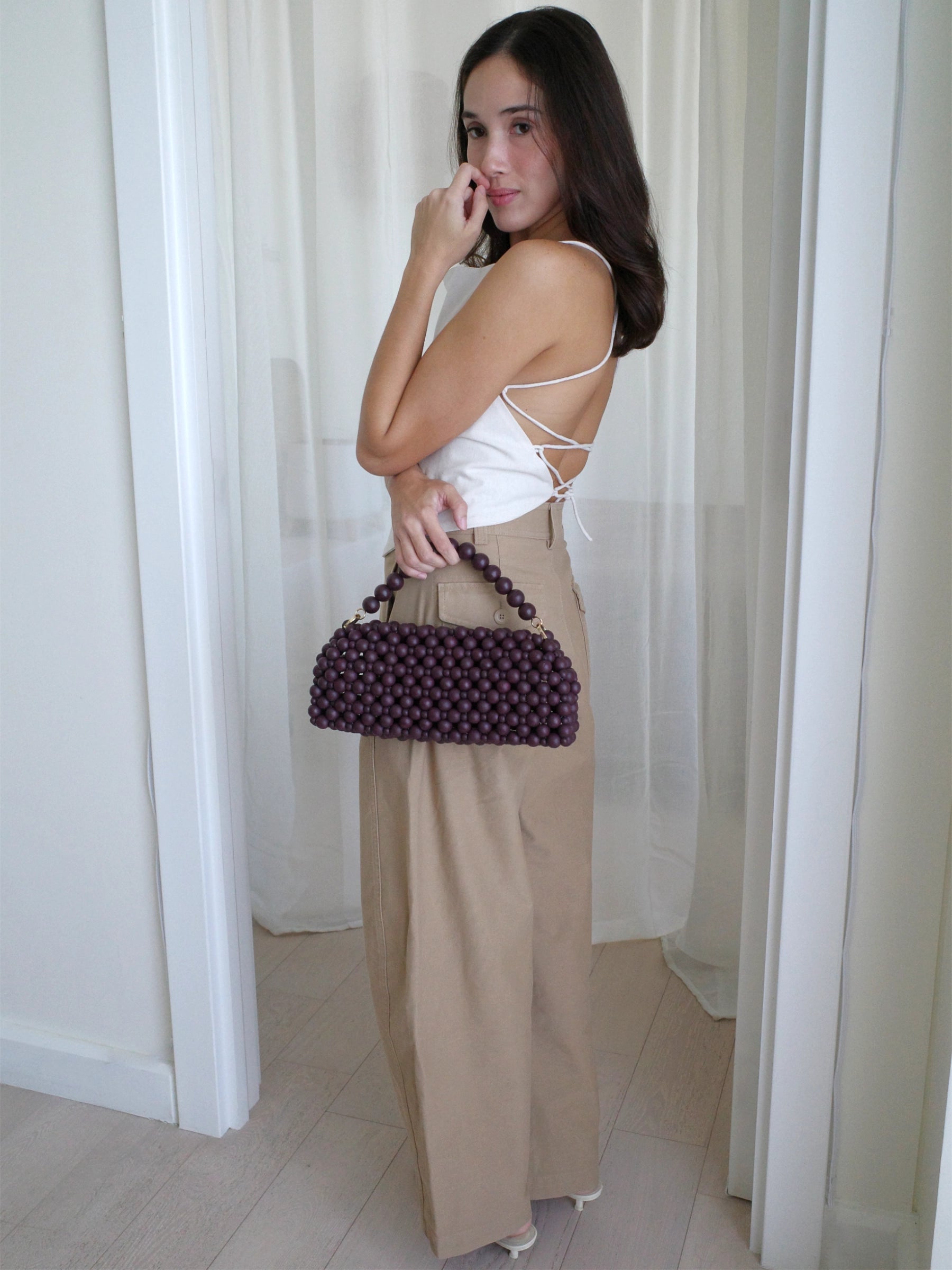 A woman stands in a doorway wearing a white strappy tank top and beige wide-leg trousers, holding a burgundy  beaded handbag.