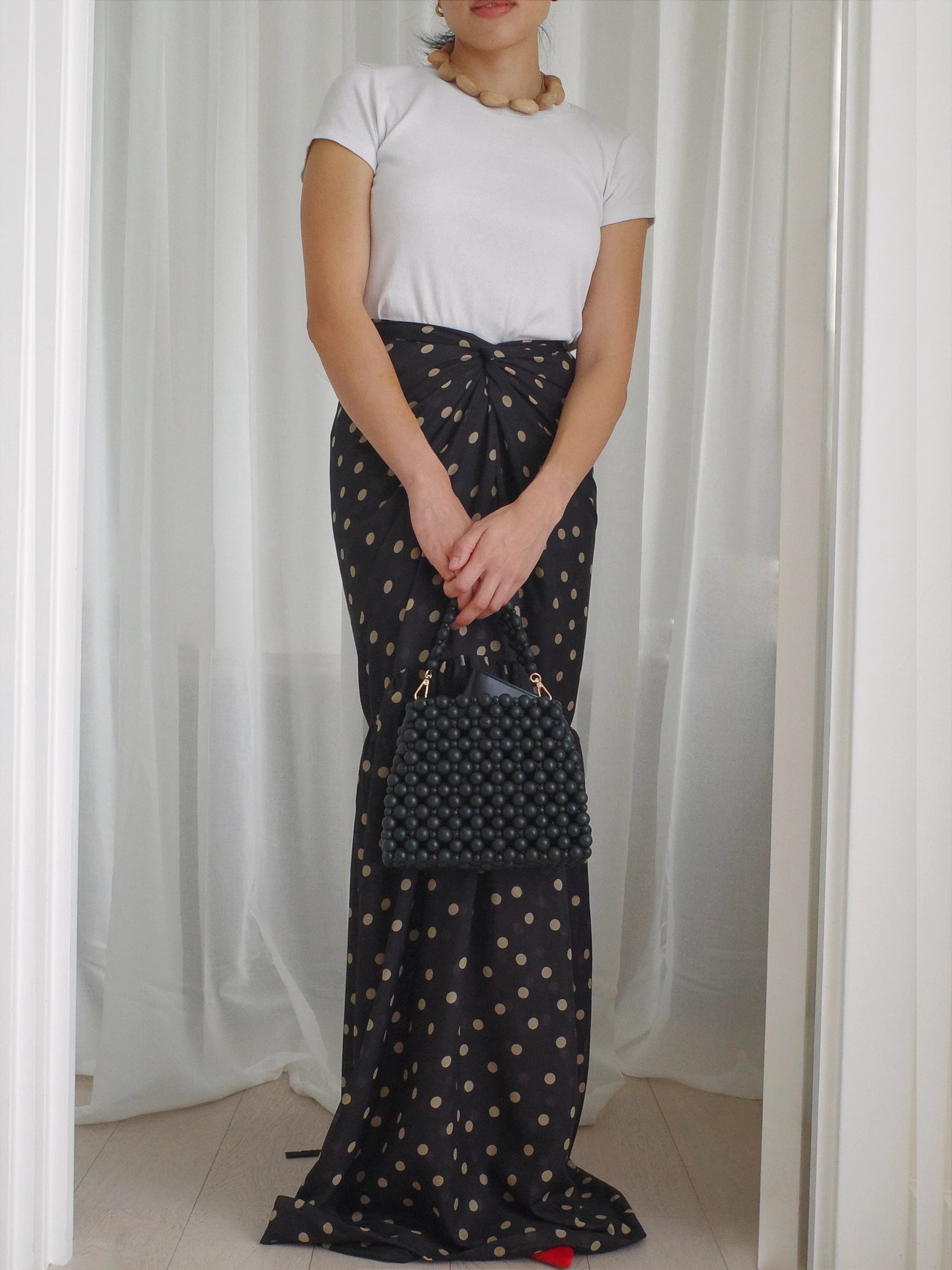 Person wearing a long black patterned skirt and holding a black beaded matching handbag against a white curtain background.