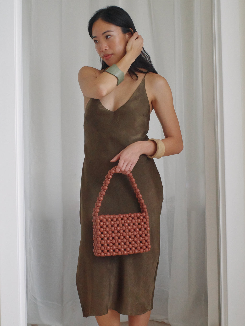 Woman holding a pink beaded handbag wearing a brown dress against a white curtain background